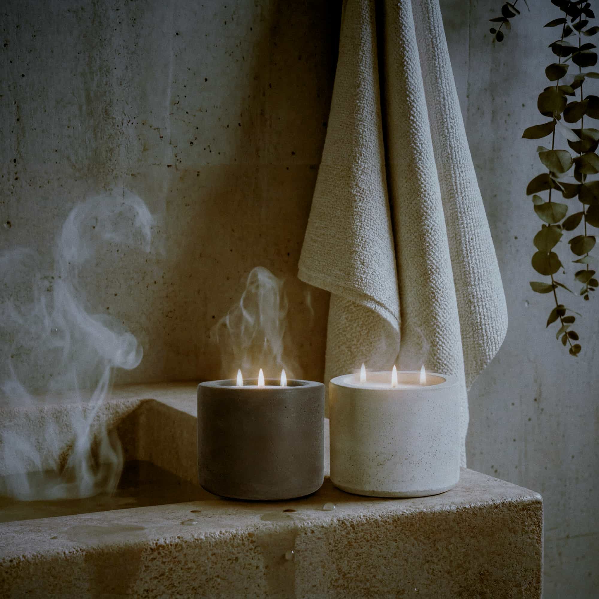 A white concrete candle jar lit with three wicks, and a black concrete candle jar also lit with three wicks on the ledge of a sandstone ledge with steam coming from the bath and a cream colored bath towel hanging on the wall behind.