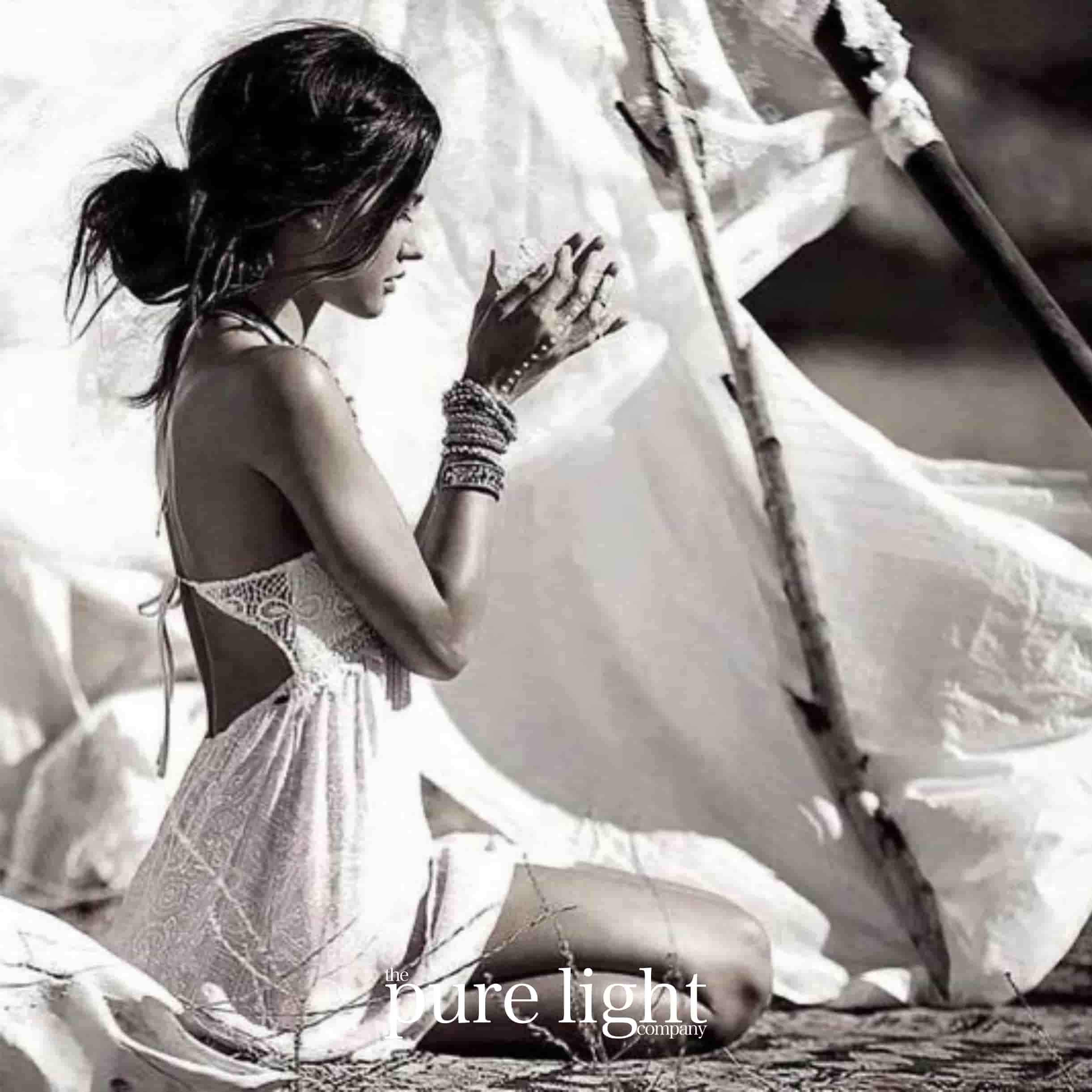 A pretty woman kneeling in a white, open-backed boho-style dress, with her hands pressed together and adorned with bracelets with The Pure Light Company logo for the Boho Chick Collection of all-natural home fragrance.