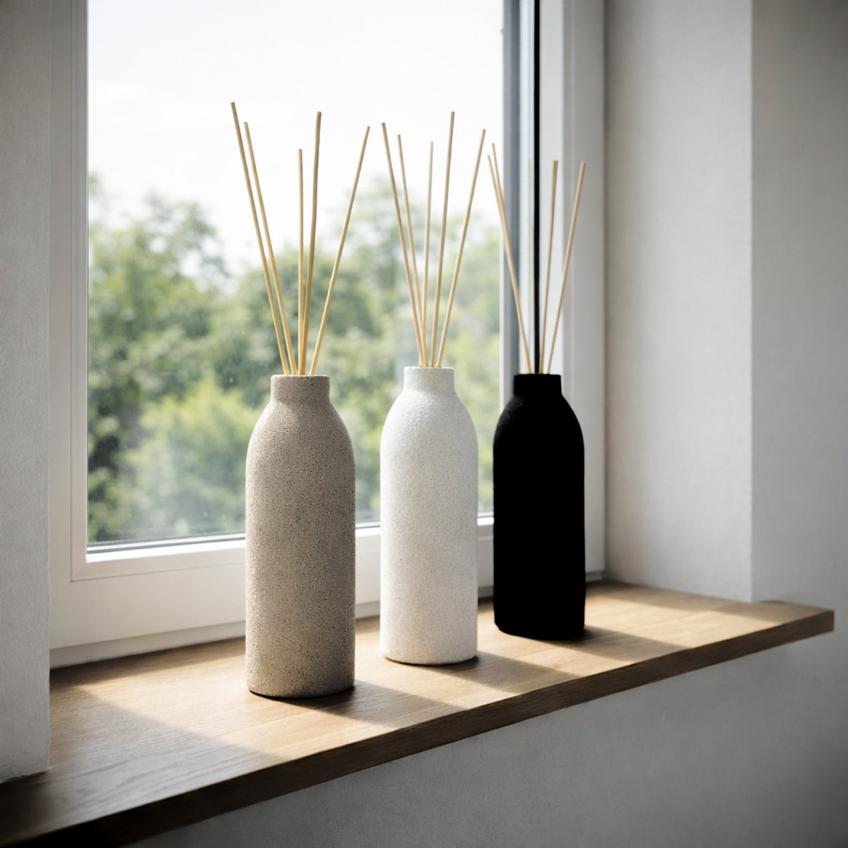 Three concrete diffusers by The Pure Light Company with reed diffusers on a windowsill with a view of greenery outside.