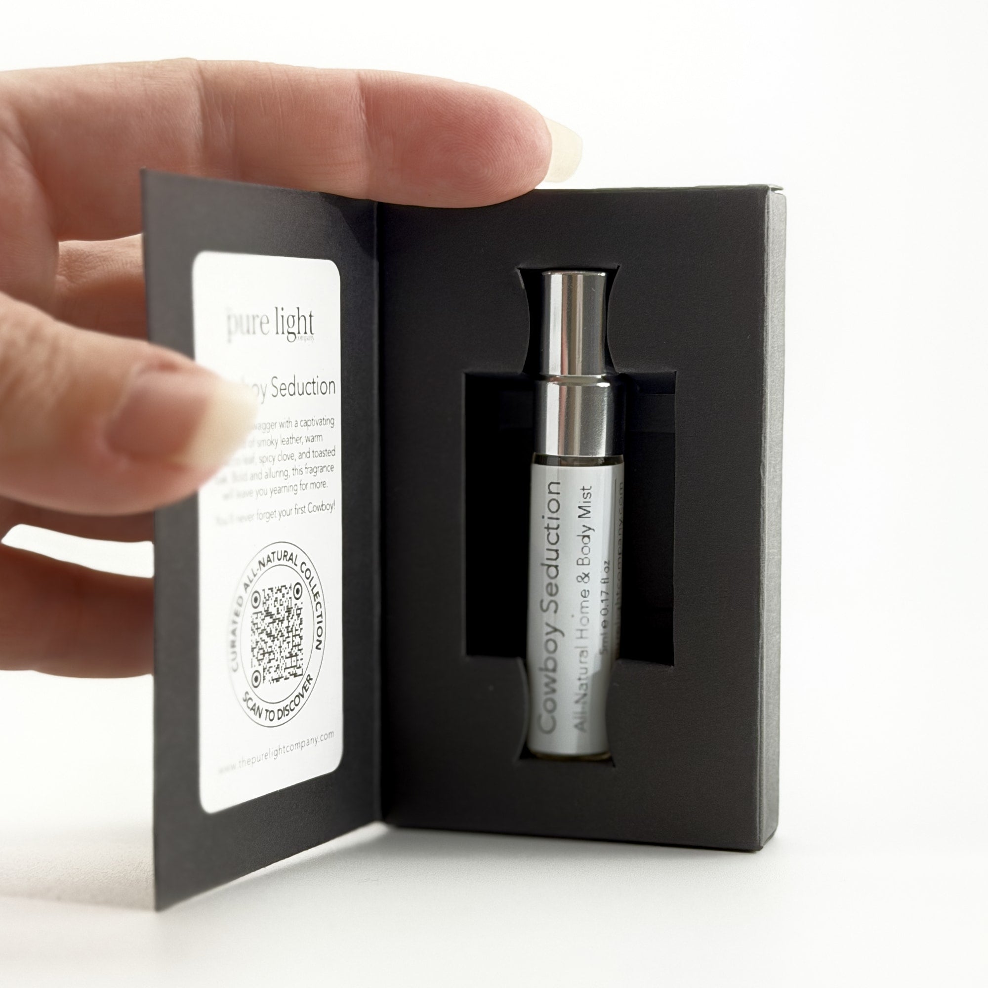 Hand holding a black box with a Cowboy Seduction 5ml Home & Body Mist bottle inside, on a white background by The Pure Light Company