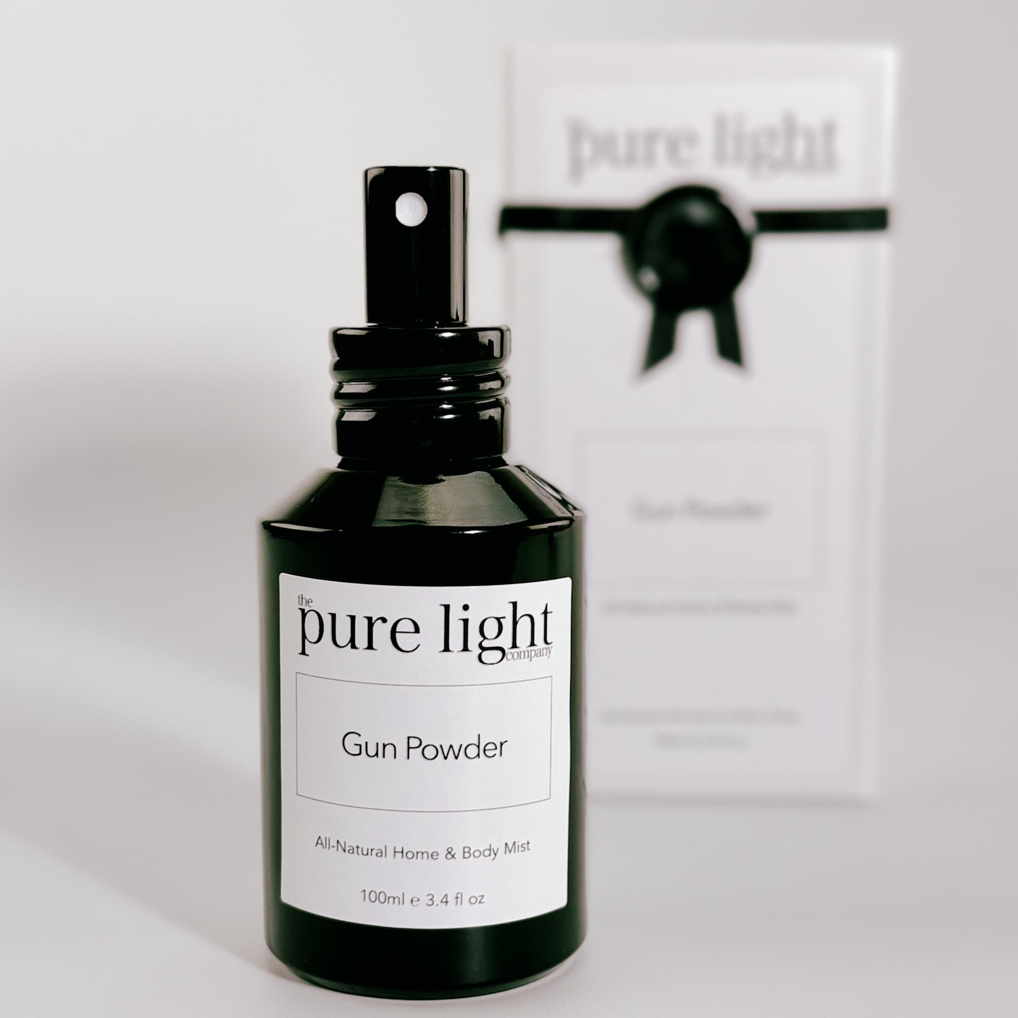 A bottle of The Pure Light Company "Gun Powder" All-Natural Home and Body Mist 100ml glossy black glass bottle with a black metal spray cap, with a white packaging box ribboned with black detailing, on a white background