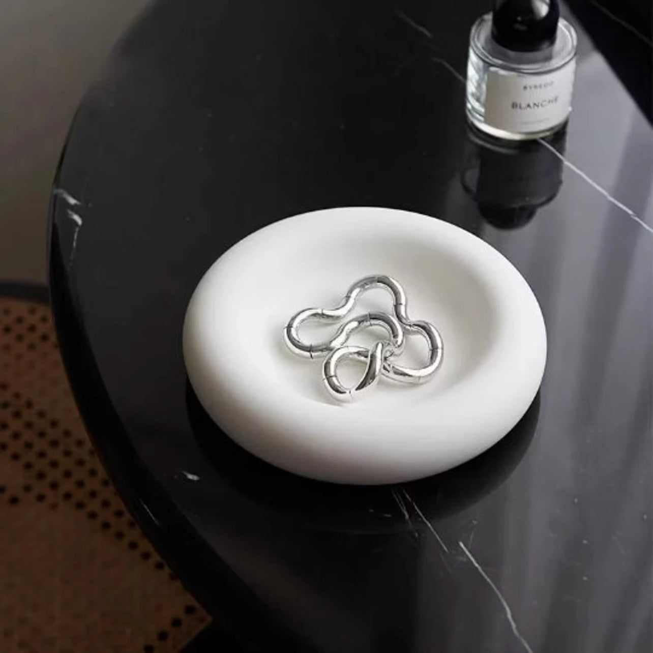 Silver necklace on a white dish with a bottle of perfume on a dark surface, branded 'the pure light company'.