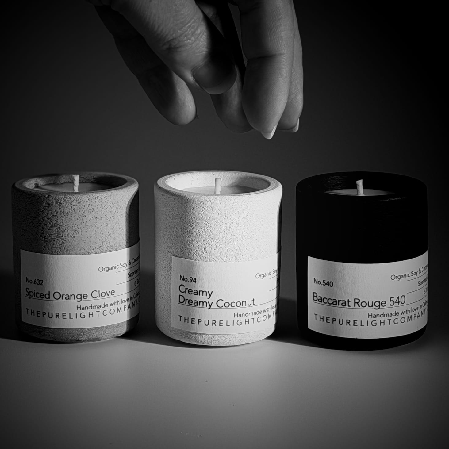 Three Mini Concrete Candles by The Pure Light Company, set against a dark background with a hand reaching to pick one up. A white jar with Creamy Dreamy Coconut, a gray jar with Spiced Orange Close and a black jar with Baccarat Rouge.