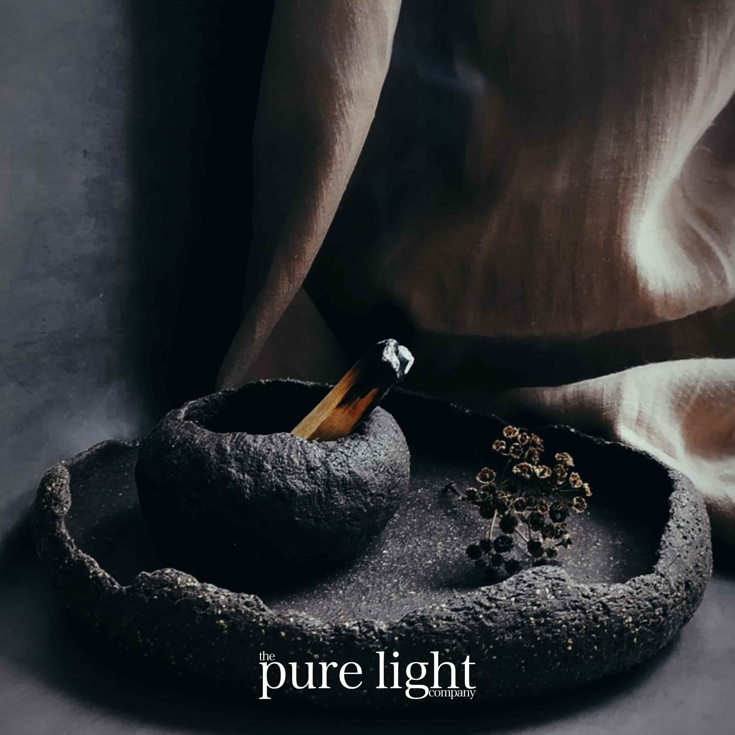 An image of an Oud Wood stick smoldering on a rustic tray with The Pure Light logo.