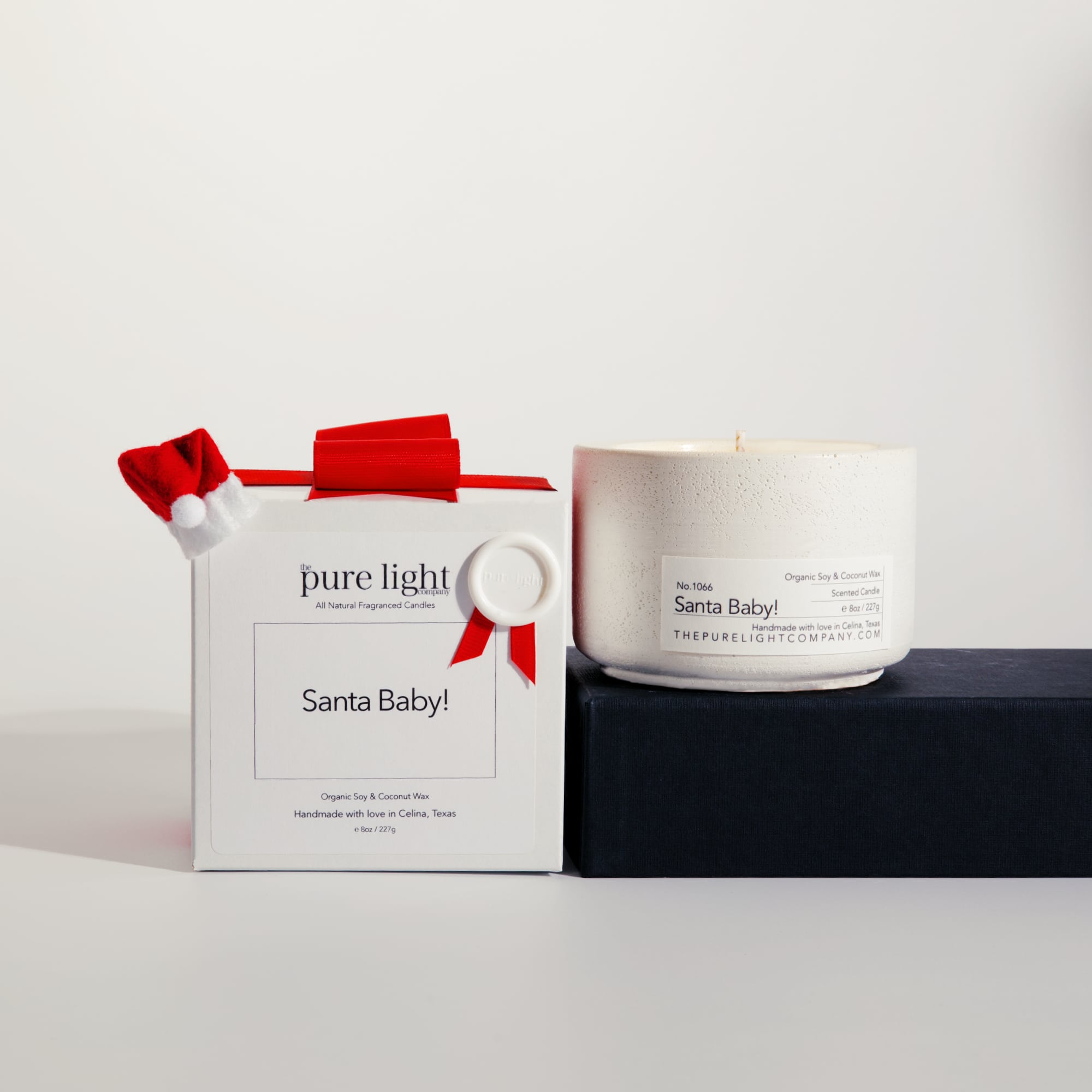Medium Santa Baby! white concrete candle on a prop beside a gift box with ribbon and wax seal by The Pure Light Company.
