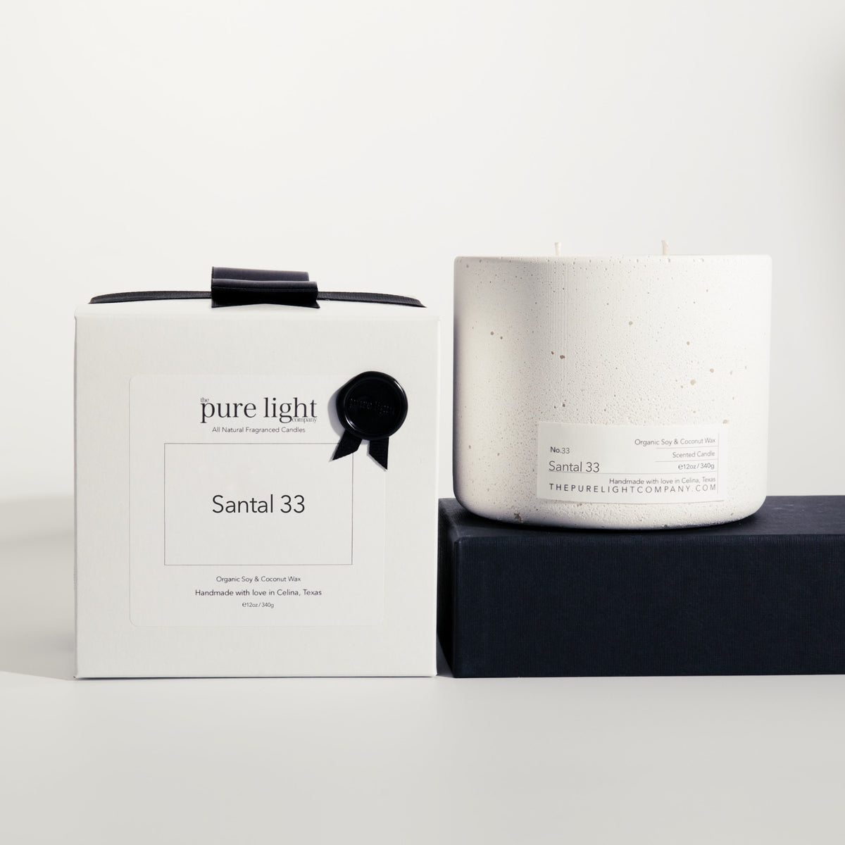A white concrete candle jar next to its white box packaging, both labeled 'Santal 33' by 'The Pure Light Company' with a black ribbon detail, set against a clean white background with a dark pedestal.