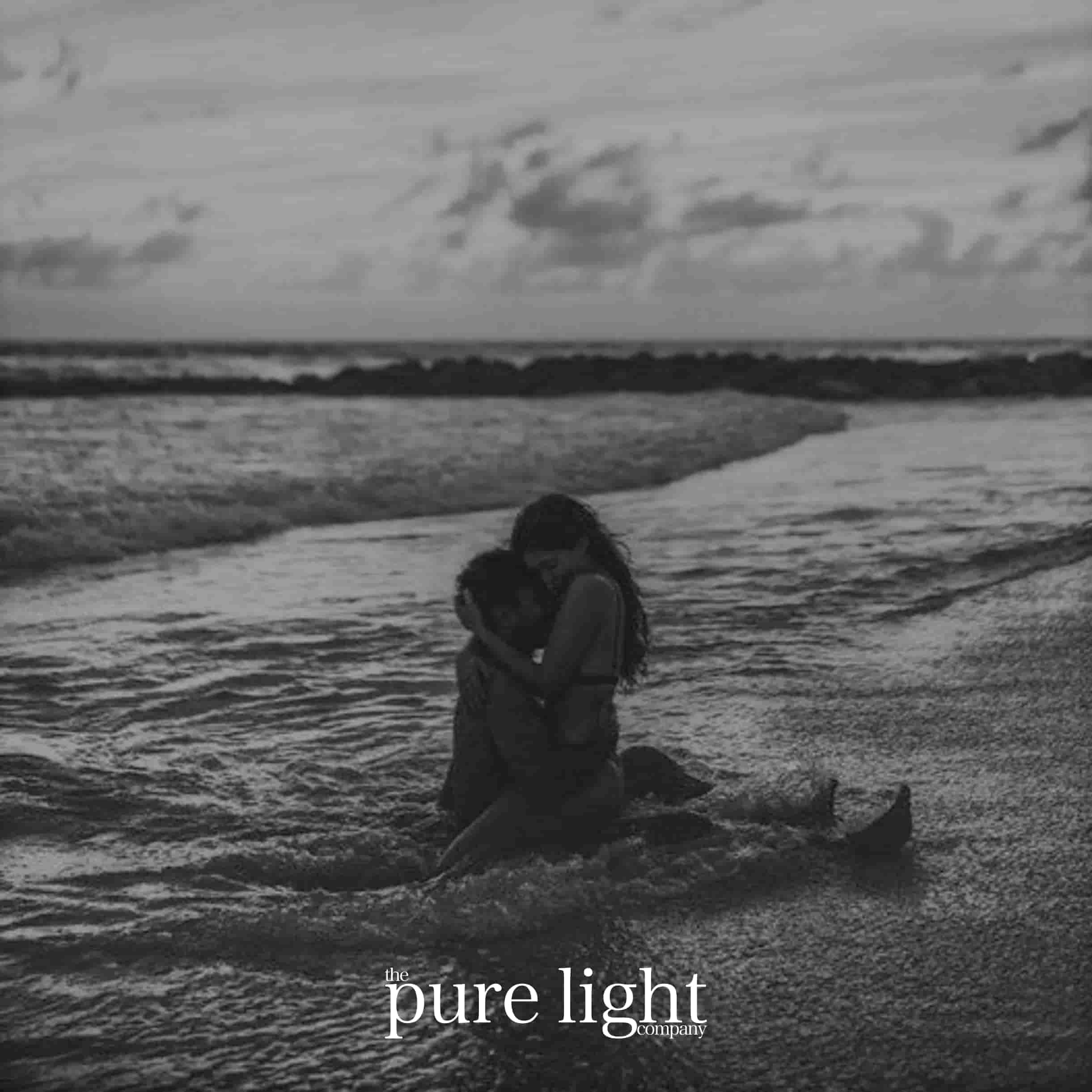 A black and white image of a couple embraced on a beach with waves crashing around them, with The Pure Light Company logo for the Sex On The Beach collection of all-natural home fragrance
