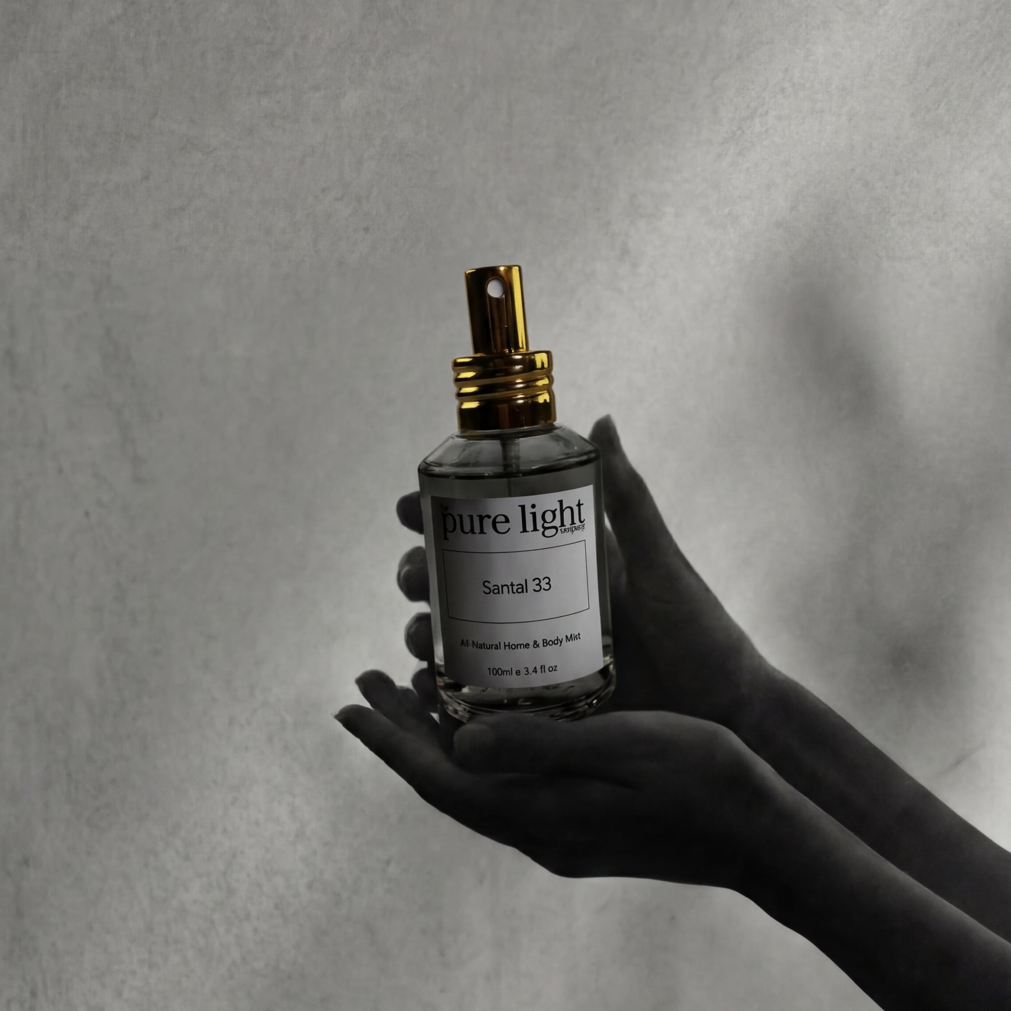 A dark moody image of hands holding up a bottle of Santal 33 in a transparent glass spray bottle with a gold metal spray head, by The Pure Light Company