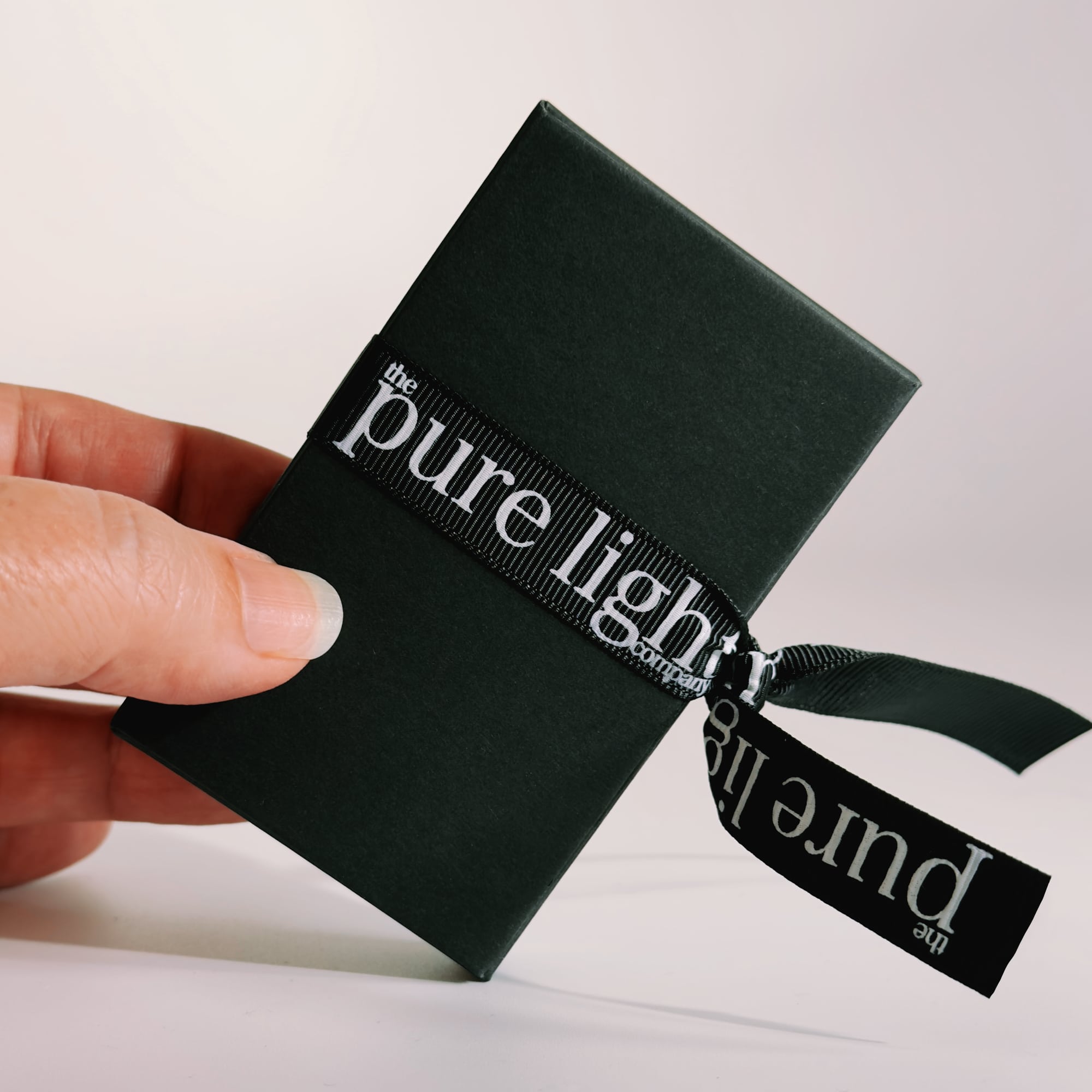 A hand holding a black rectangular box tied with a black ribbon printed with the white text 'the pure light company' all-natural home and body mist 5ml.