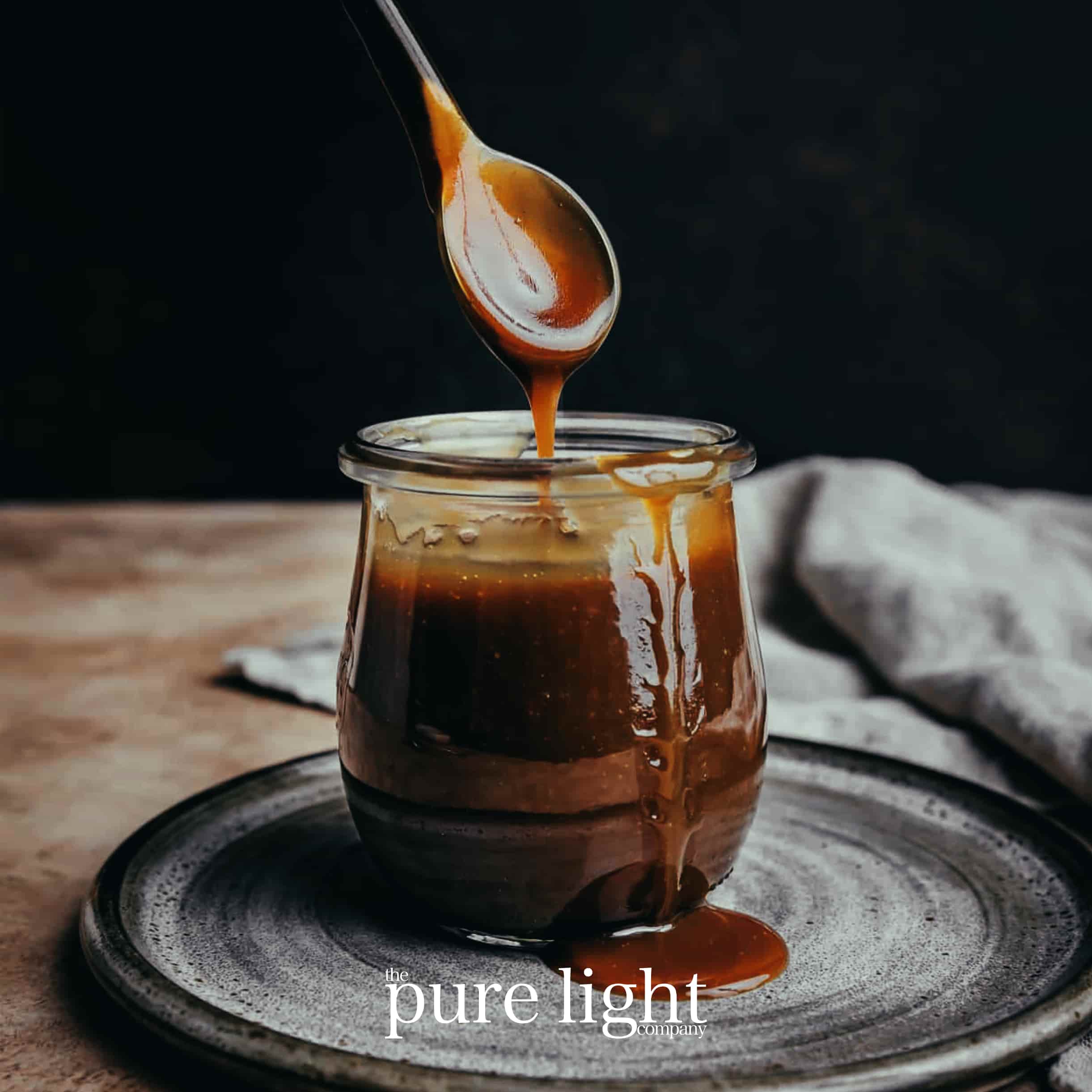 A mouthwatering image of a jar of butterscotch sauce, with a spoon dripping the sauce onto a plate with The Pure Light Company logo for the Comforting Butterscotch collection of all-natural home fragrance.