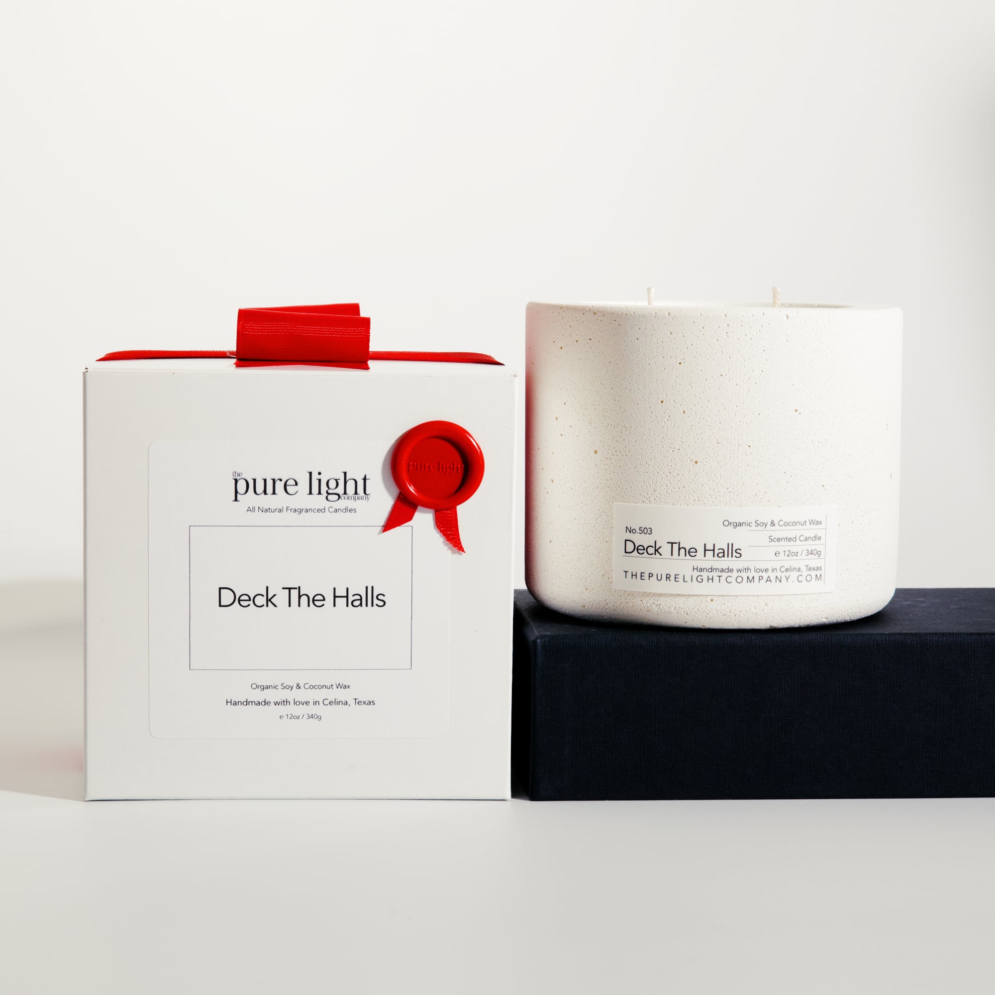 Large Deck The Halls white concrete candle on a prop beside a gift box with ribbon and wax seal by The Pure Light Company