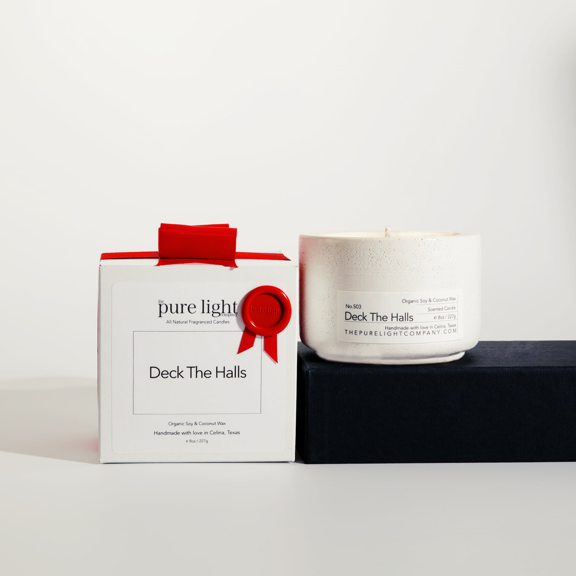 Medium Deck The Halls white concrete candle on a prop beside a gift box with ribbon and wax seal by The Pure Light Company.