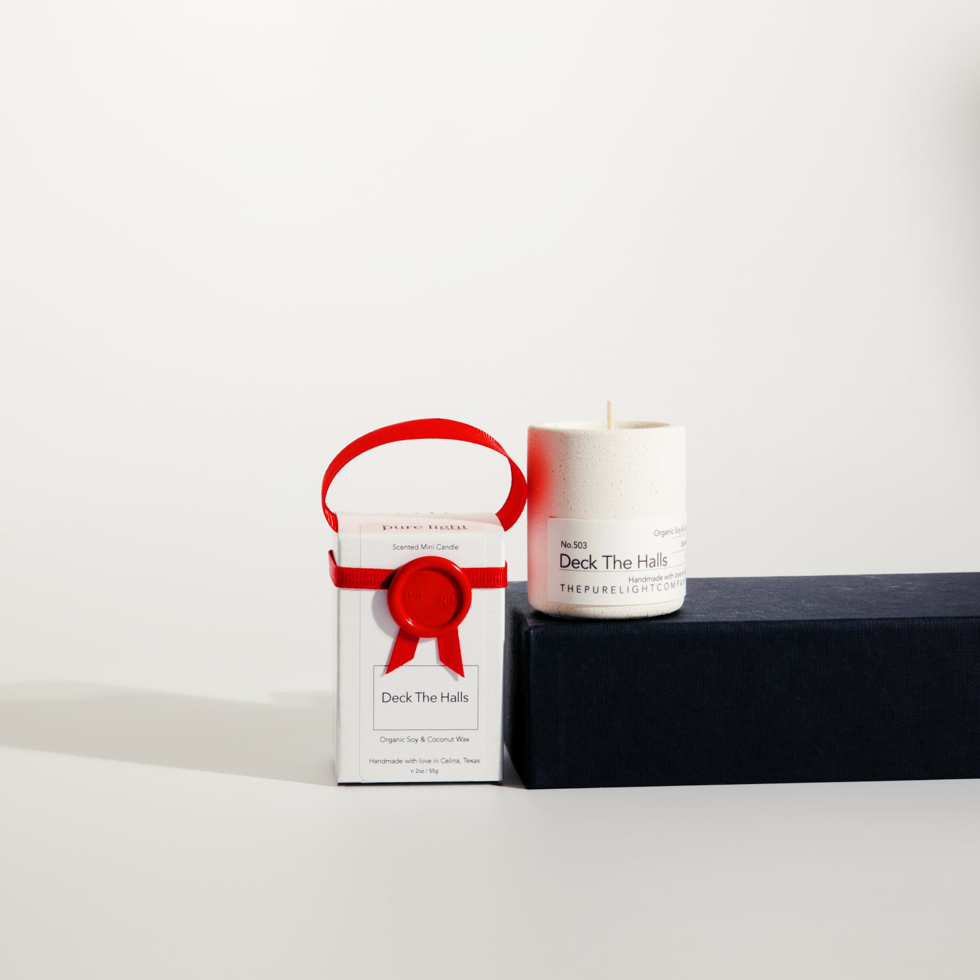 Mini Deck The Halls white concrete candle on a prop beside a gift box with ribbon and wax seal by The Pure Light Company.