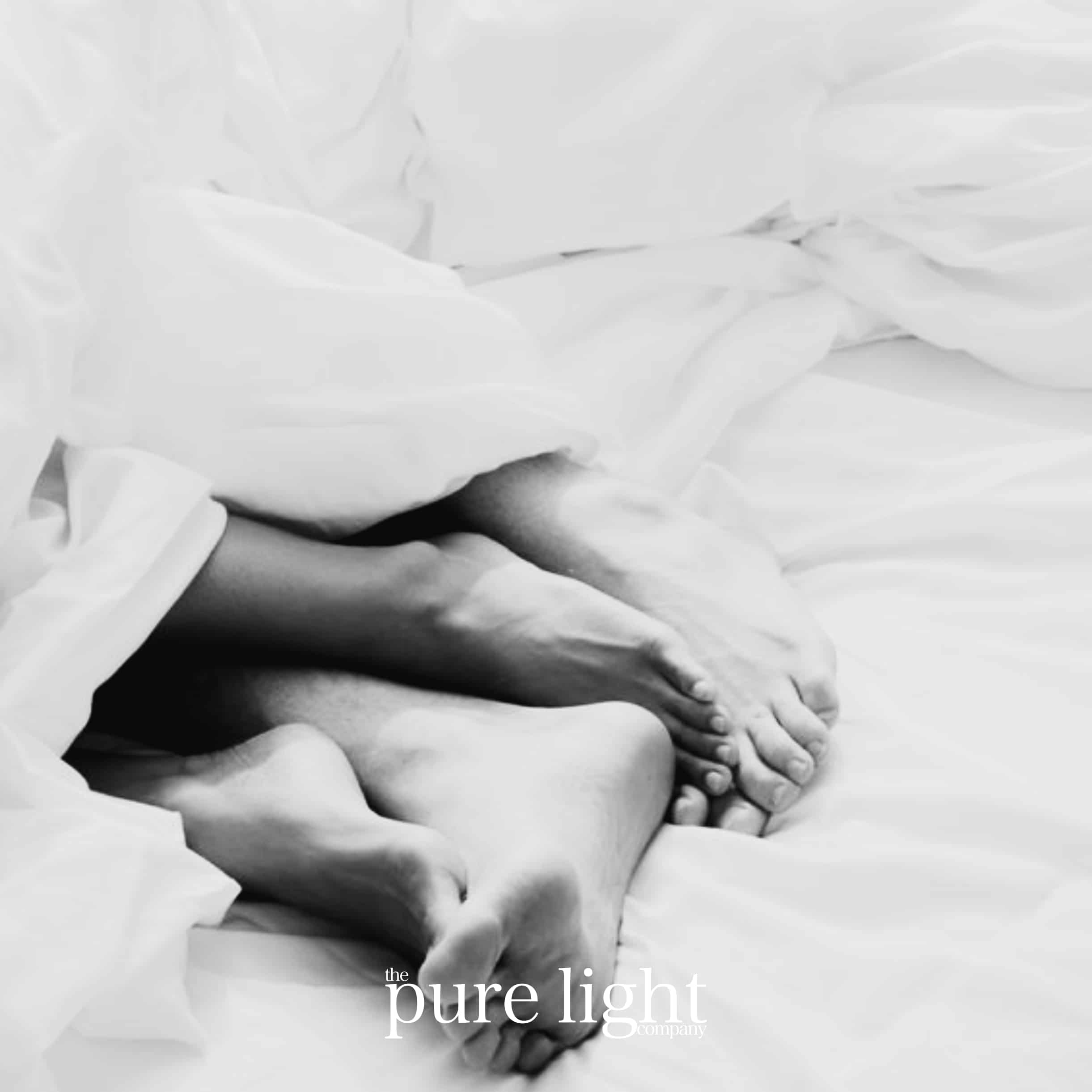 Black and white image of a couple's feet snuggled together in clean, crisp bedding with the Pure Light Company branding