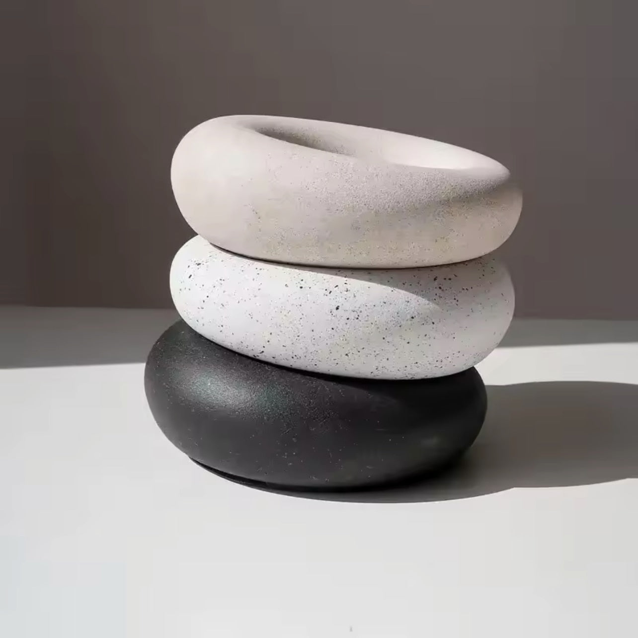 Stack of three concrete trinket dishes in black, white terrazzo, and sandstone on a neutral background by The Pure Light Company