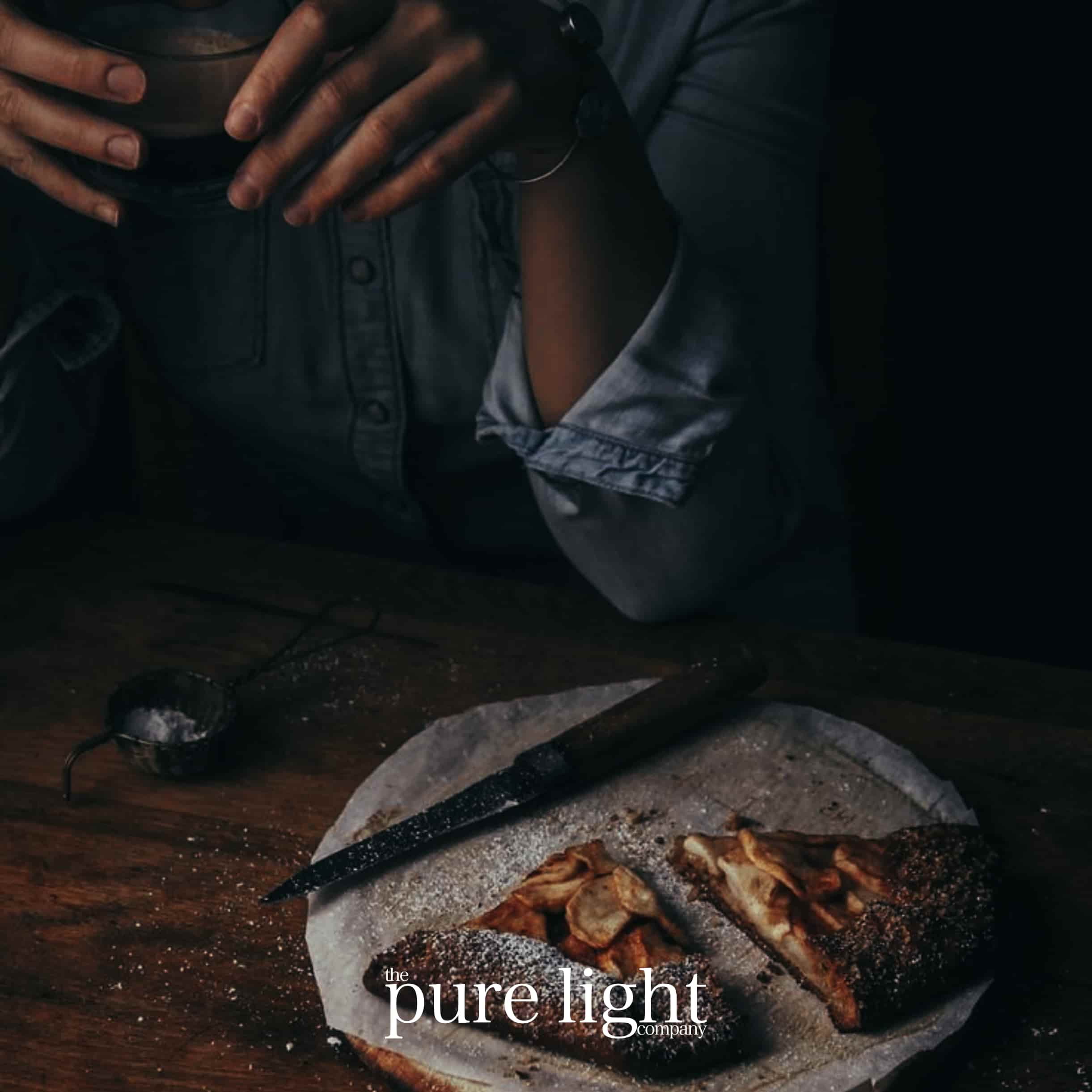 A close-up image of delicious Hot Apple Pie coated in powdered sugar next to a kitchen knife and The Pure Light Company logo.