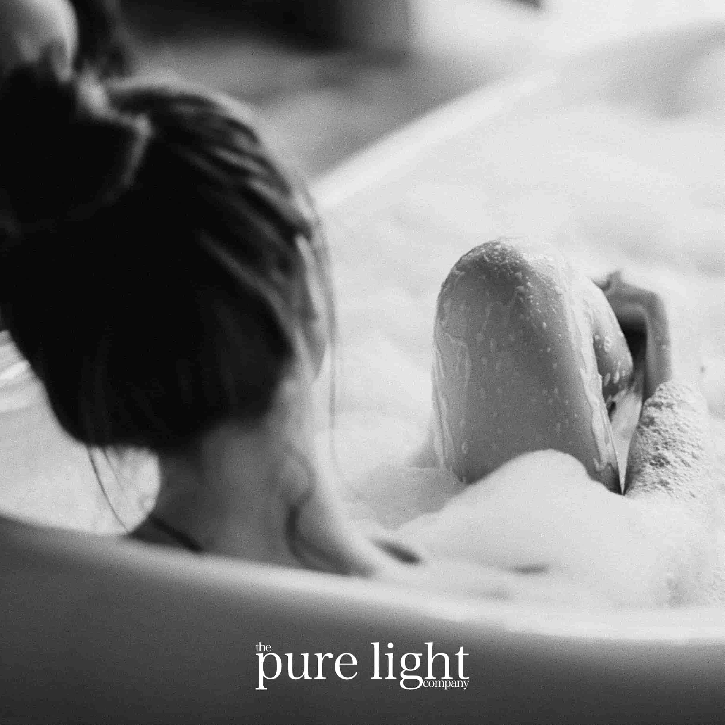 A black and white image of a person in a bathtub filled with bubbles with her hair tied back with The Pure Light Company logo for the Me Time all-natural home fragrance collection.
