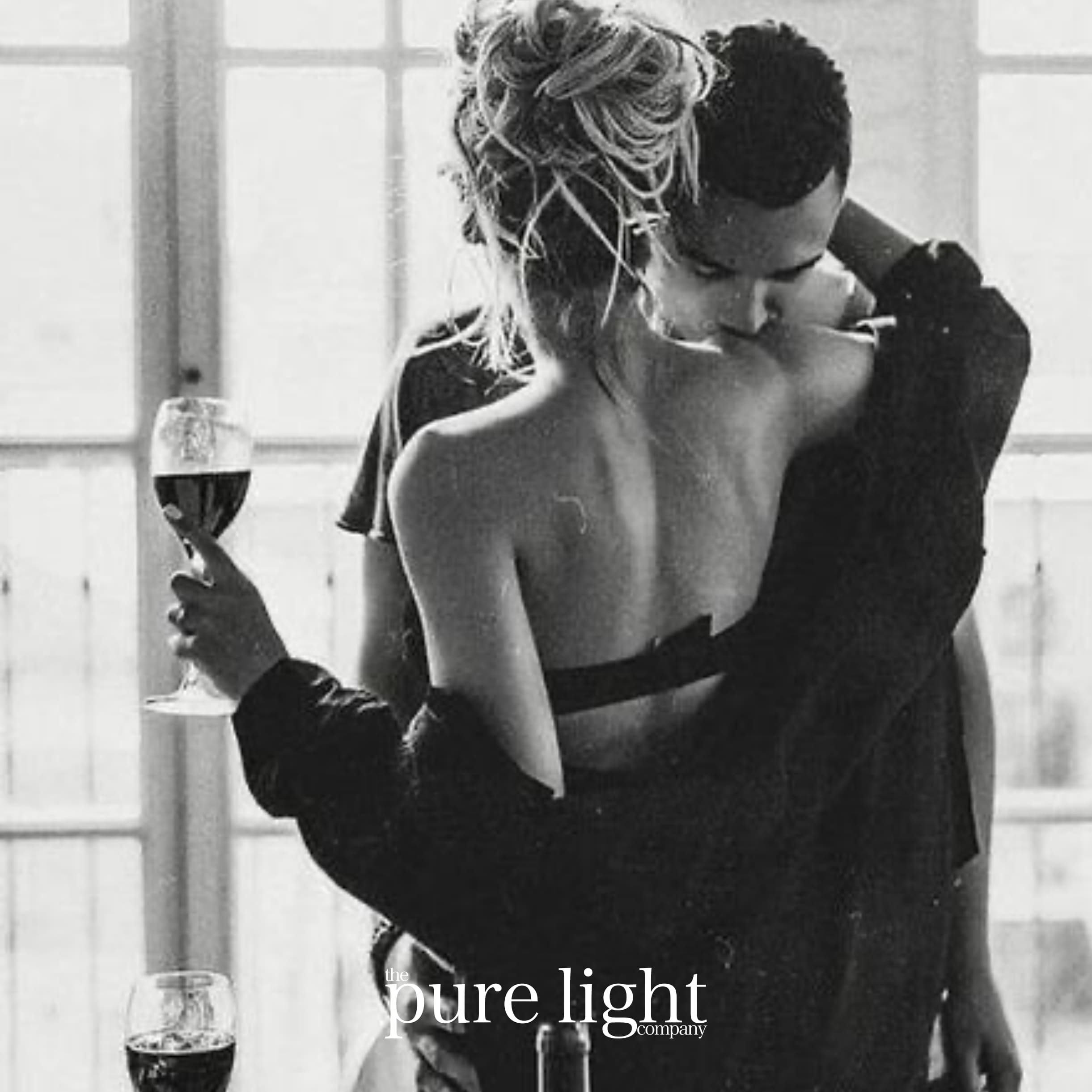 A black and white photograph of a couple embracing, with one person holding a glass of wine. The background is dimly lit and The Pure Light Company logo for their Red Red Wine all-natural fragranced home collection.