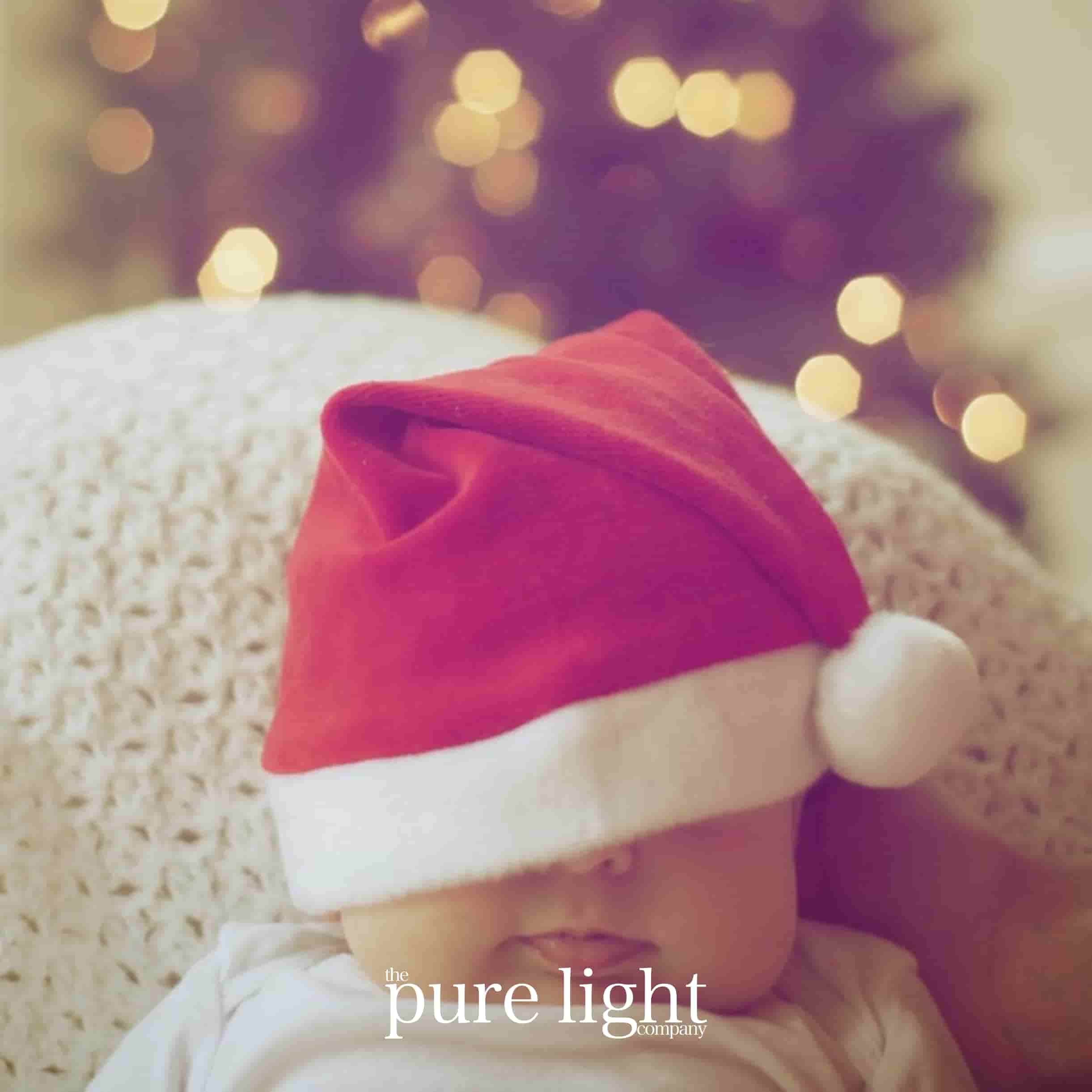 Baby wearing a red Santa hat with blurred lights in the background for The Pure Light Company's Santa Baby! all-natural home fragrance 