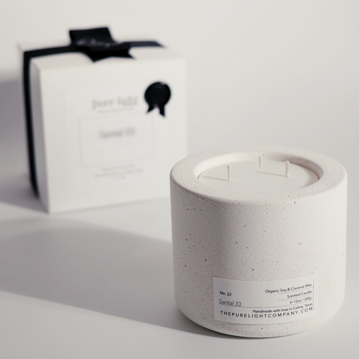 A white concrete candle jar with a speckled off-white finish and a white label, shown in front of its matching white and black branded box on a clean white background saying 'Santal 33' by 'The Pure Light Company'