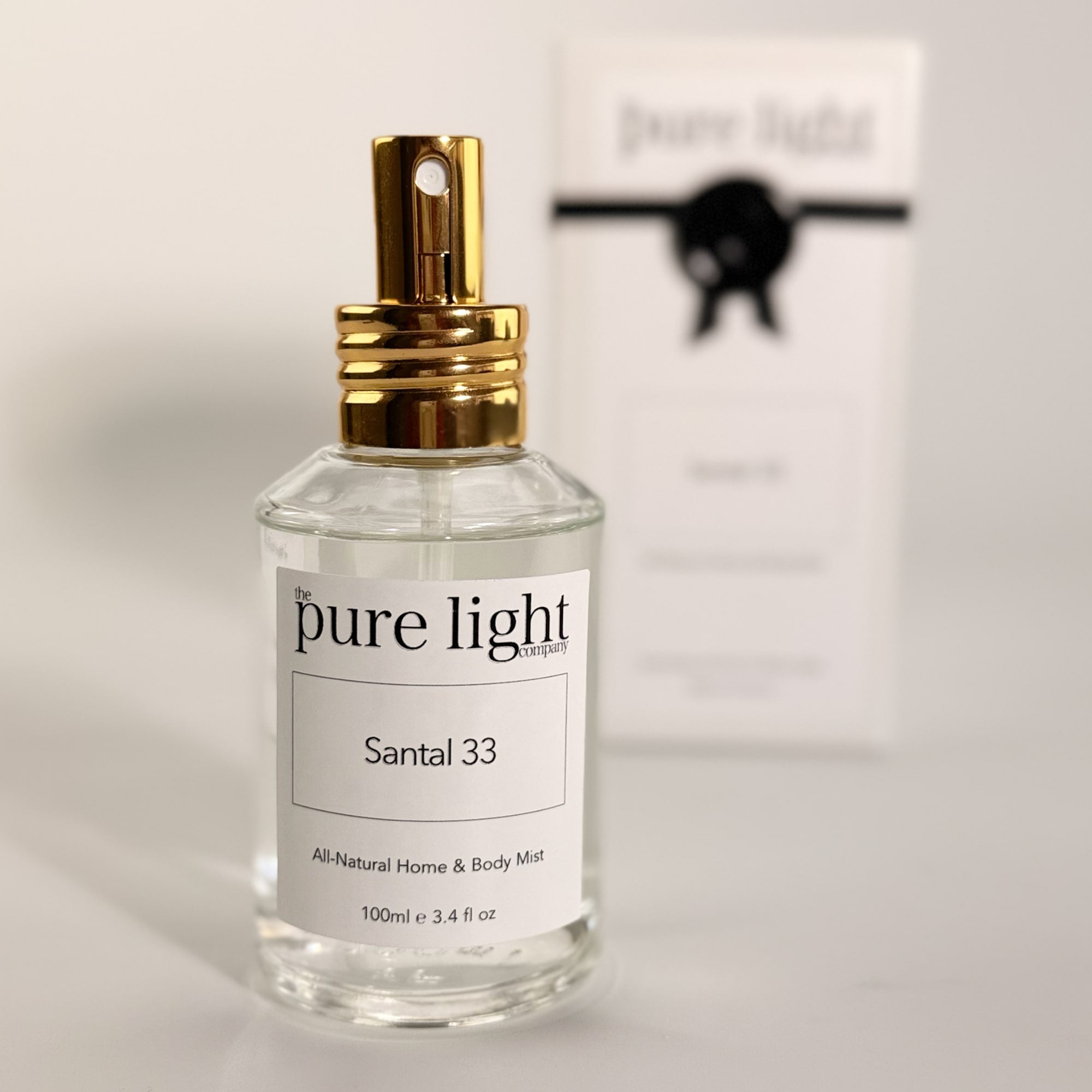 Bottle of The Pure Light Company Santal 33 LeLabo Dupe All-Natural Home and Body Mist 100ml clear glass bottle with a gold cap, and it's packaging on a white background.
