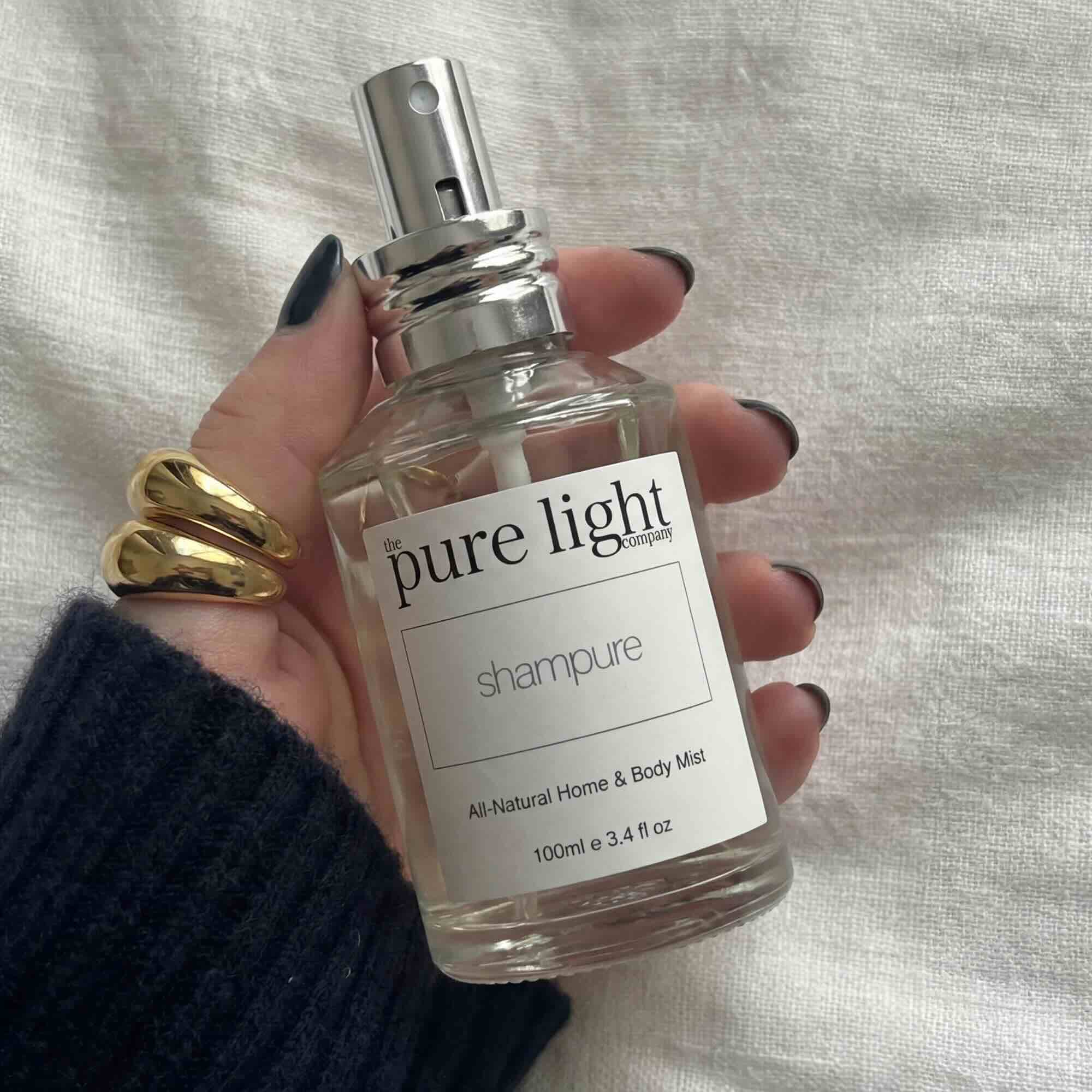 Hand with black nail polish holding a clear 100ml bottle of Shampure All-Natural Home & Body Mist by The Pure Light Company with a silver spray cap, against a linen cloth background