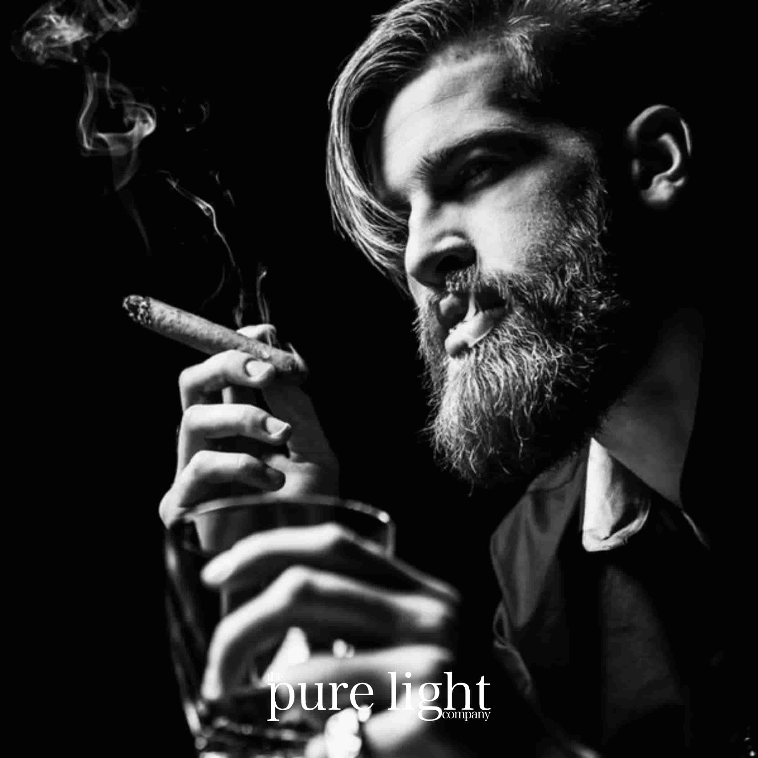 A black and white image of a sultry bearded man smoking a cigar holding a glass of bourbon with The Pure Light Company logo.