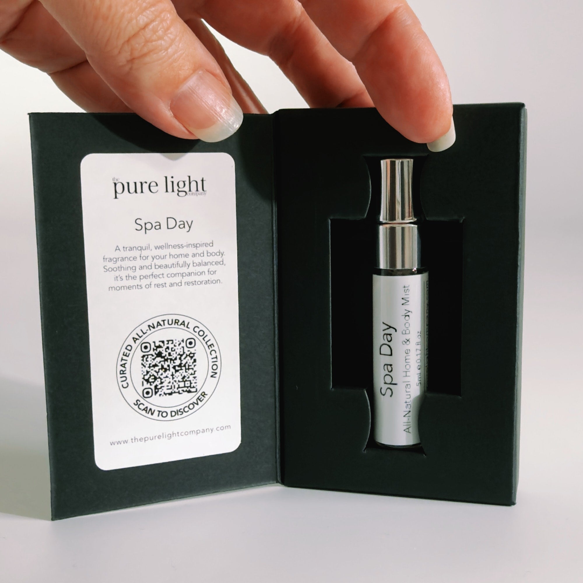 A hand holds a black presentation box containing a small white and silver spray bottle labeled 'Spa Day' by the pure light company. The left side of the box has a white label with product details and a QR code.
