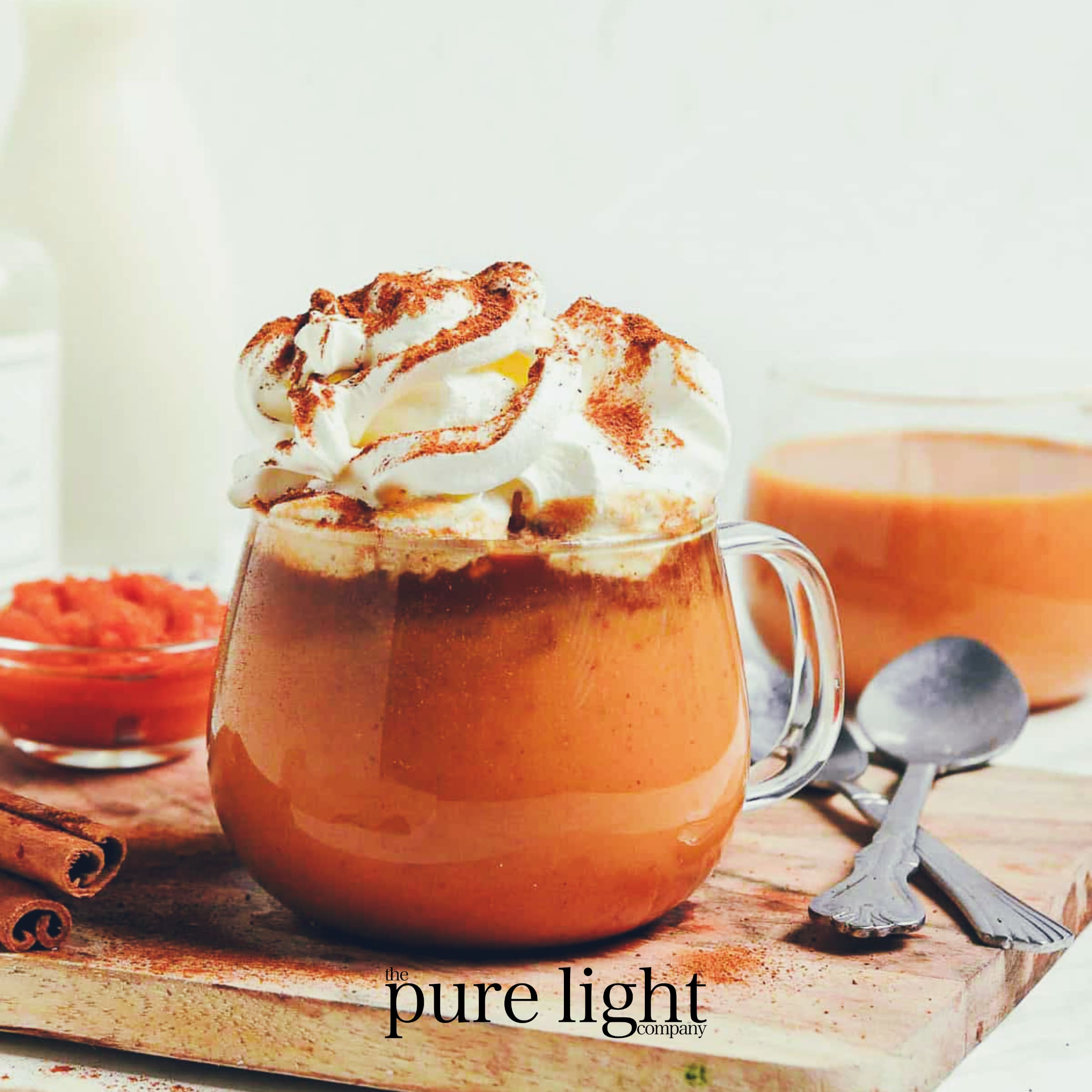 A glass mug filled with a delicious spiced pumpkin latte brimming with whipped cream and a sprinkle cinnamon and The Pure Light Company logo.